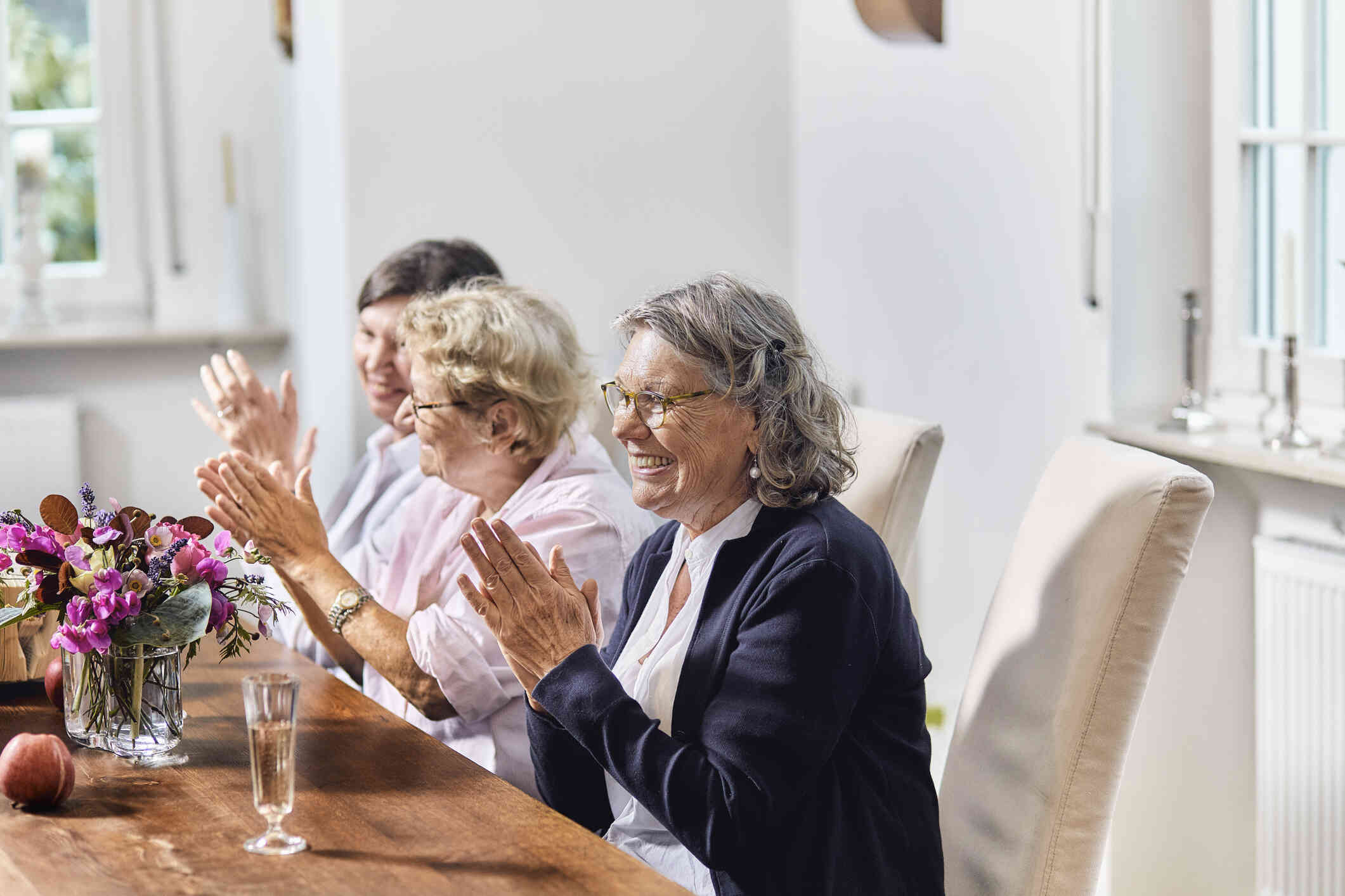 A group of elderly woman sit at a dining room table together while clapping and  smiling.